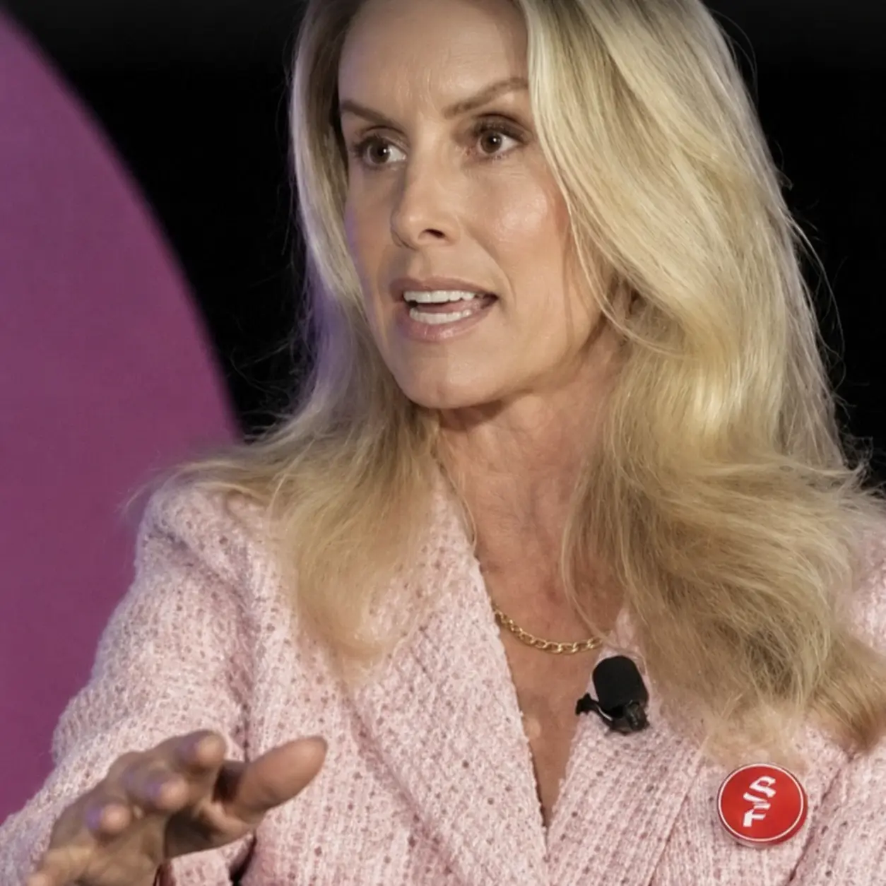 League One Volleyball's owner speaking on stage in a pink jacket, wearing a round red Rodela badge on her lapel.